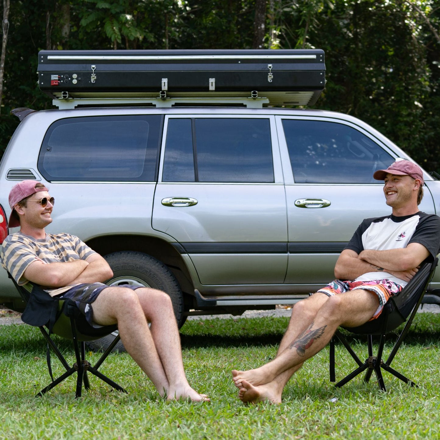 Two men sitting on Ozzi Gear chairs in front of a silver SUV with a roof rack.