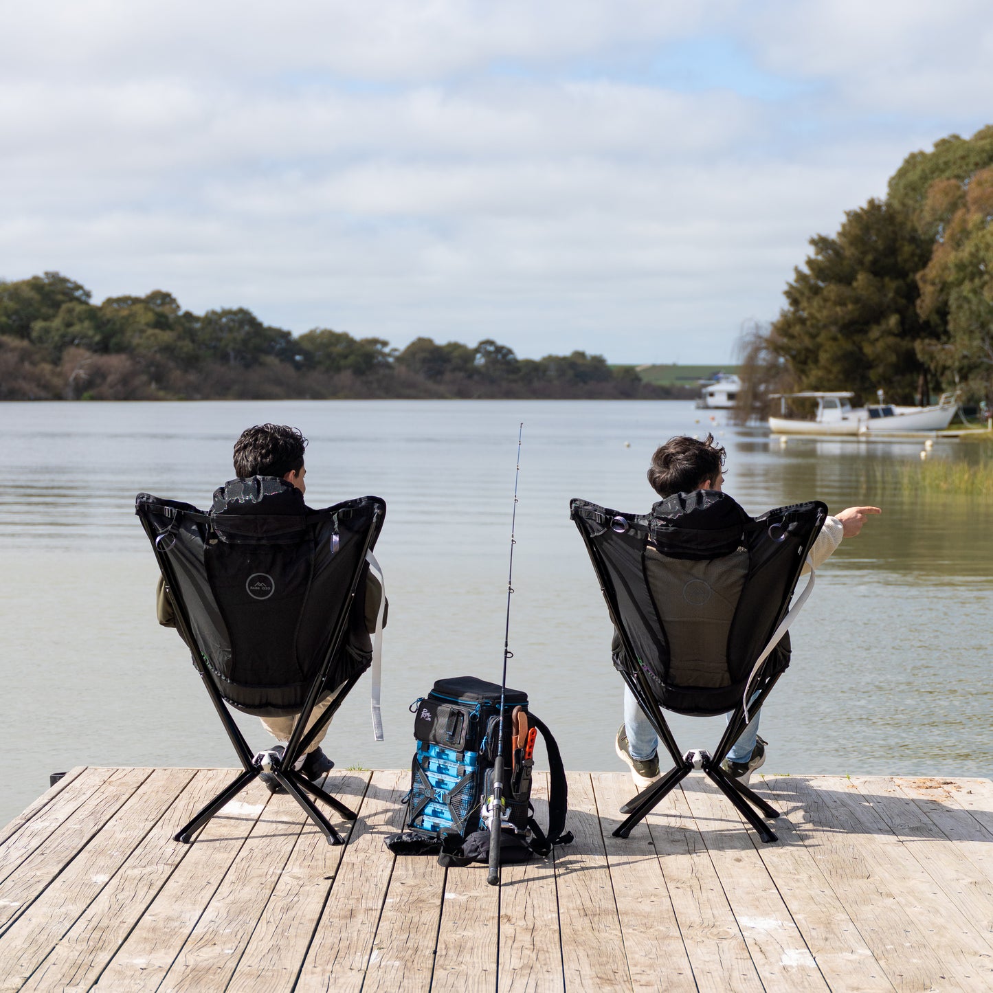 Two people sitting on the foldable Ozzi gear lounger chairs by a lake, fishing.