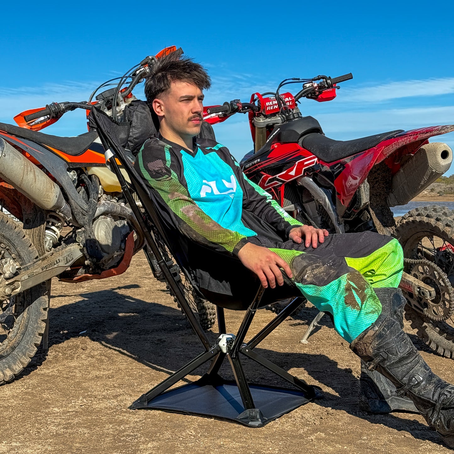 A man sitting on an Ozzi Gear lounger next to off-road vehicles with a clear blue sky.